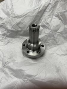 A Metal Spindle With A Threaded Top And Multiple Bolt Holes At The Base, Resembling Components From Battlebots, Sits On Crumpled White Paper.