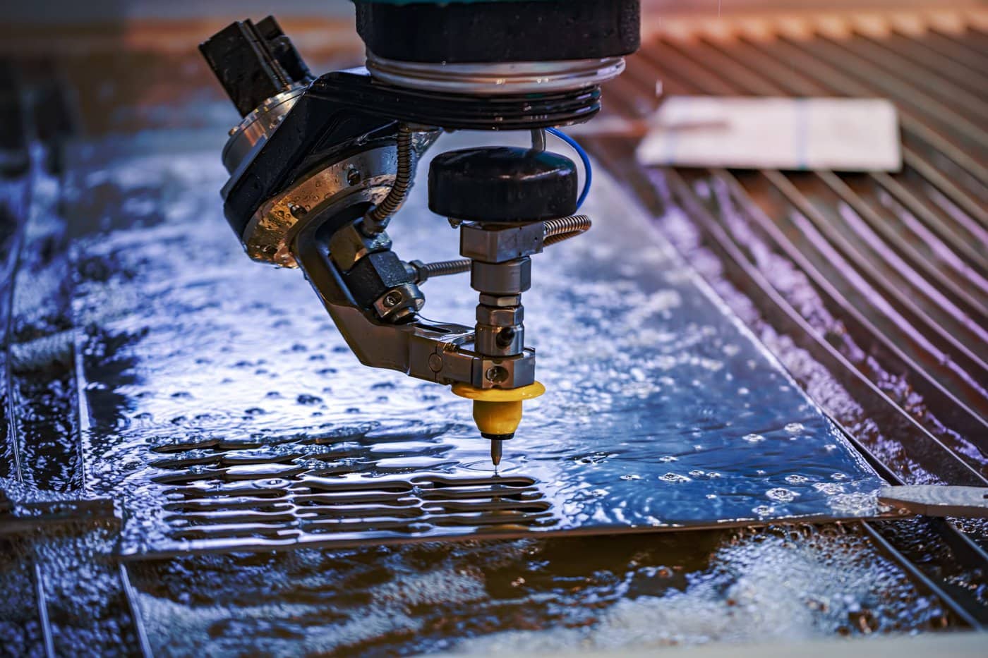 A close-up of a water jet cutting machine slicing through a metal sheet. Water sprays out from the nozzle under high pressure, creating precise cuts. The surface is wet, with small water droplets scattered around.