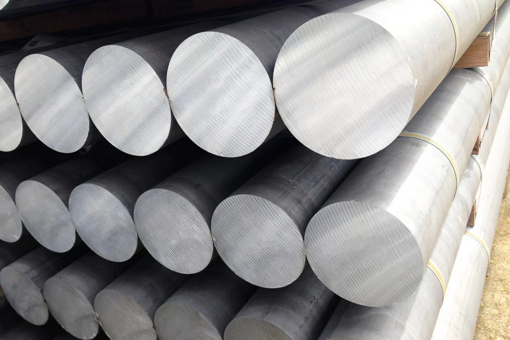 Stacks Of Large Cylindrical Metal Rods Are Piled Horizontally In Two Rows. The Rods Have A Shiny, Smooth Surface And Are Arranged With Their Circular Ends Facing Outward. They Appear To Be Industrial Materials.