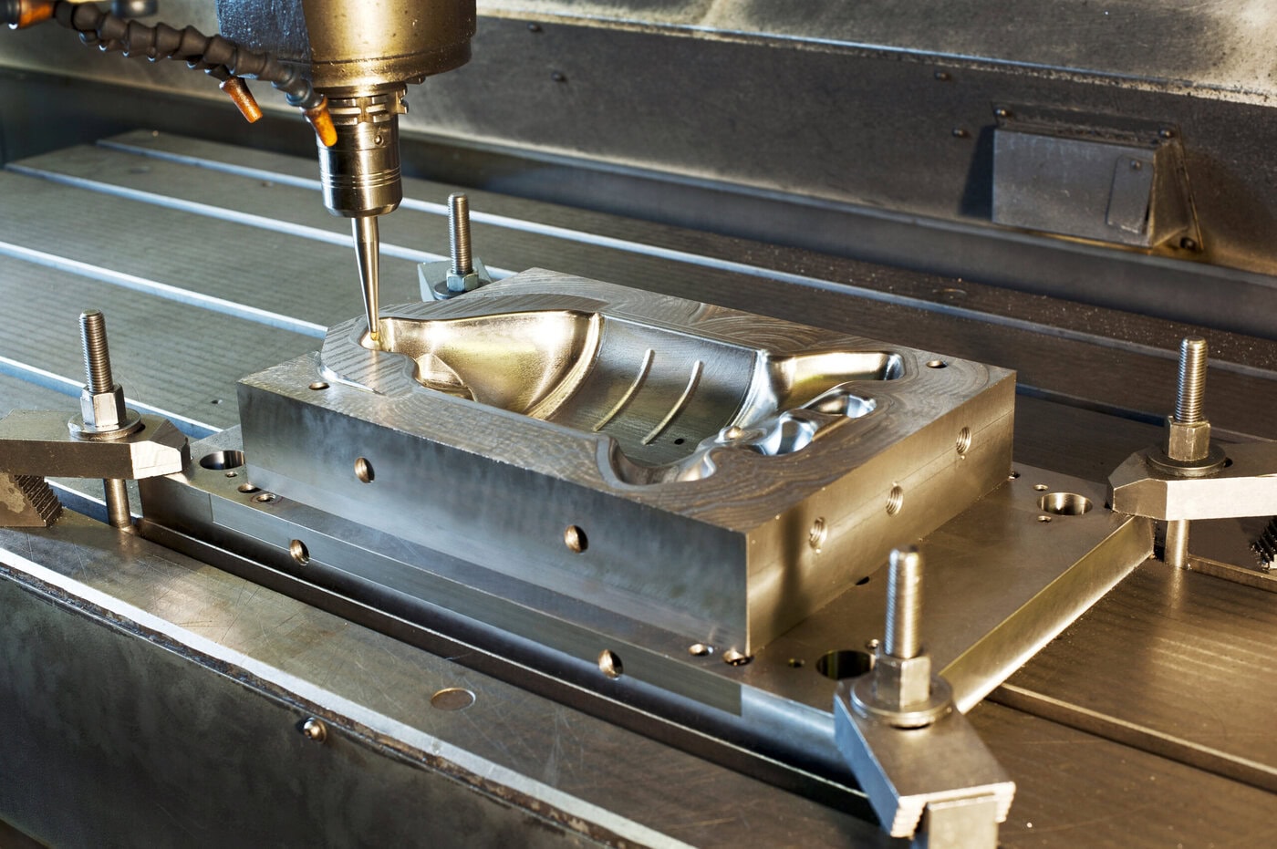 A CNC machine operates on a metal mold, with clamps securing the workpiece on the table. The cutting tool is in action, creating a detailed cavity in the workpiece, showcasing precision machining.