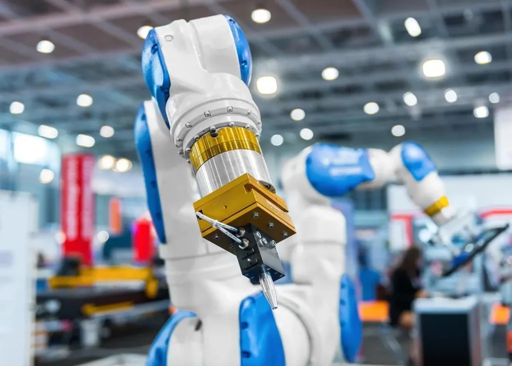 A Close-Up Of A Robotic Arm In A Factory Setting, With Blue And White Components And A Metallic Tool Attached At The End. The Background Shows Machinery And Blurry Figures, Suggesting A Busy Industrial Environment.