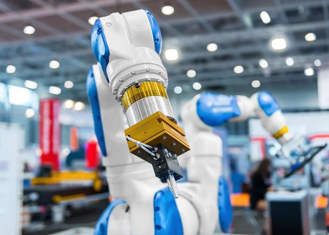 A close-up of a robotic arm in a factory setting, with blue and white components and a metallic tool attached at the end. The background shows machinery and blurry figures, suggesting a busy industrial environment.