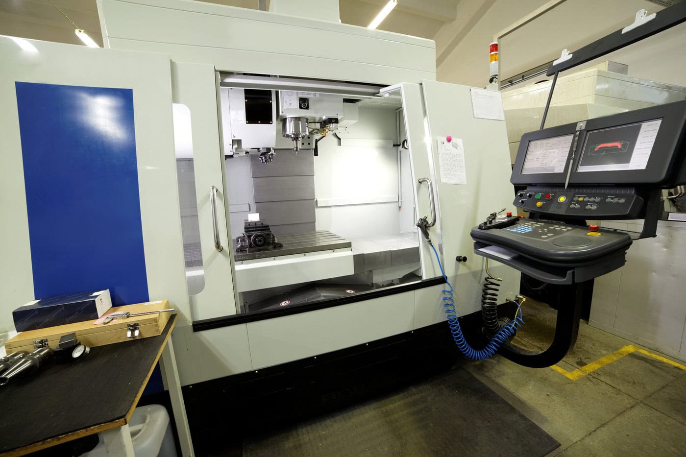 A CNC machine in a workshop is shown with its door open, revealing the interior workspace. Nearby, a control panel with multiple screens and buttons is visible. Various tools and materials are scattered on a table to the left.