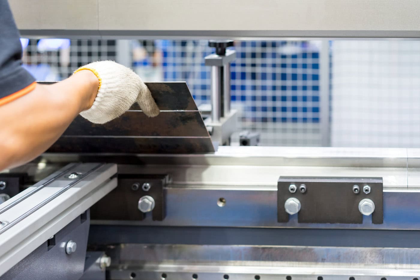 A person wearing a white glove operates a sheet metal bending machine, carefully positioning a metal sheet. The setting appears industrial, with a focus on the machinery's precision and engineering.