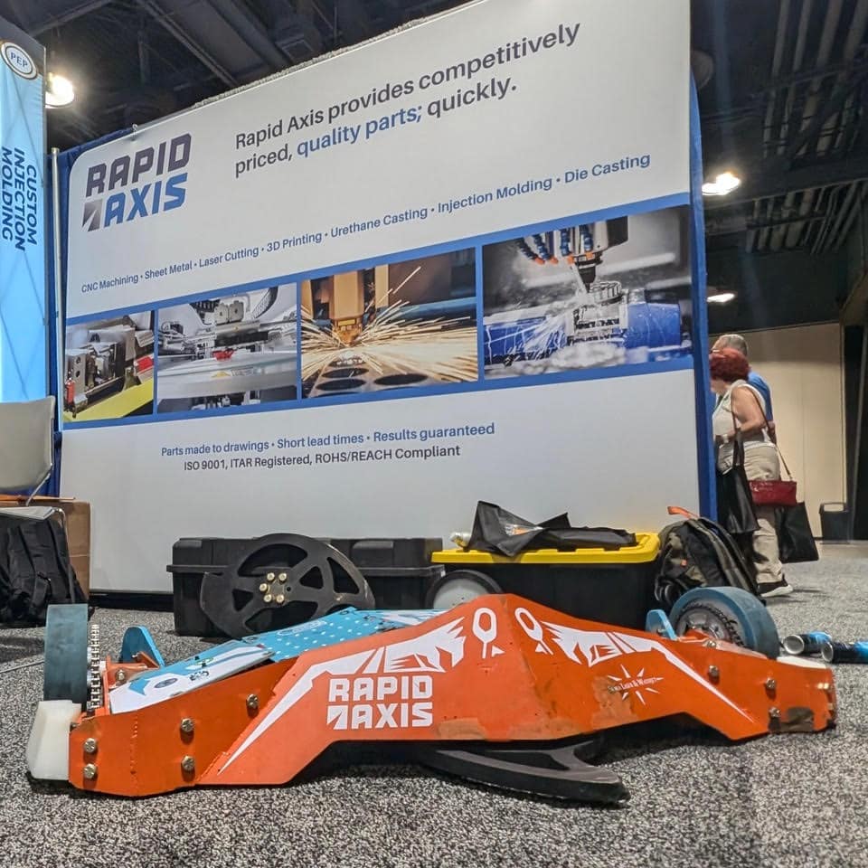 A Trade Show Booth Displays A Large Poster With &Quot;Rapid Axis&Quot; Offering Machining Services. In Front, An Orange Robotic Vehicle Resembling Those Seen In Battlebots, Adorned With The &Quot;Rapid Axis&Quot; Logo, Is Showcased On The Floor, Drawing Attention.