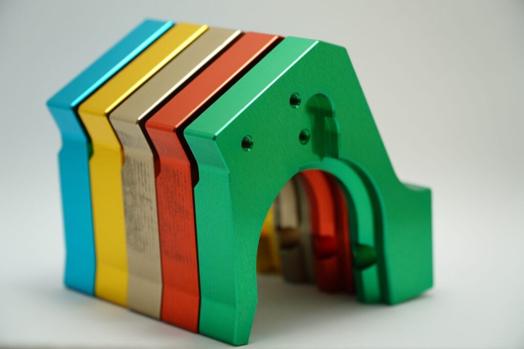 A Stack Of Colorful Wooden Blocks Shaped Like A House. The Blocks Are Arranged In A Gradient From Blue, Yellow, Metallic, Orange, To Green, Creating An Archway At The Front. The Blocks Have A Smooth, Glossy Finish.