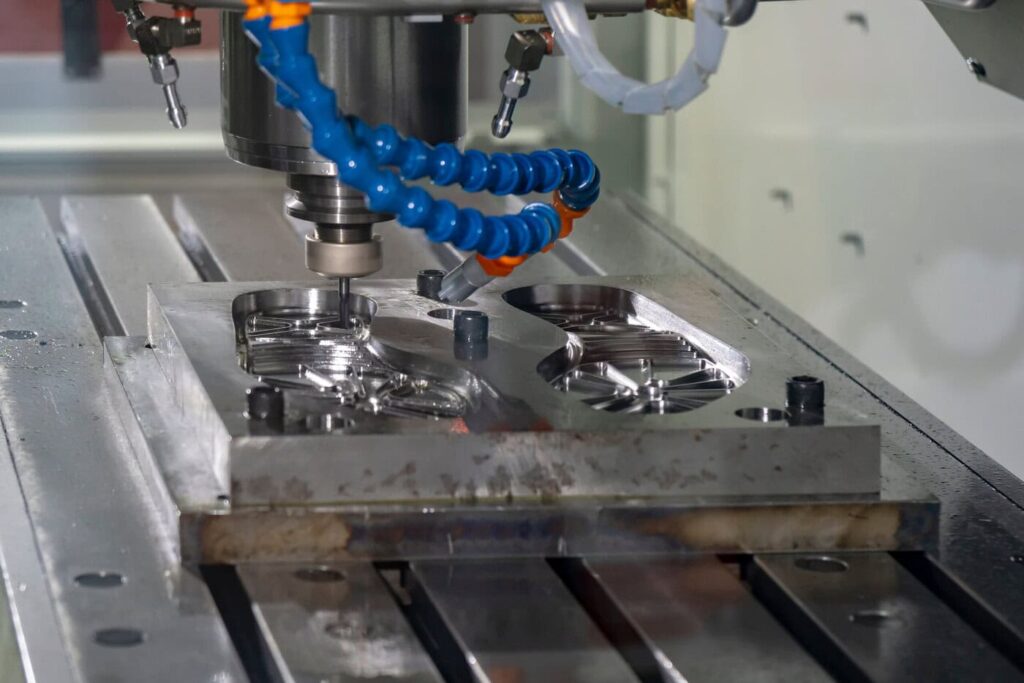 Close-Up Of A Cnc Machine In Operation, Carving Intricate Designs Into A Metal Piece. The Machine Features A Blue Coolant Hose, And Shiny Metal Chips Are Visible, Highlighting Precision Engineering And Advanced Manufacturing Technology.