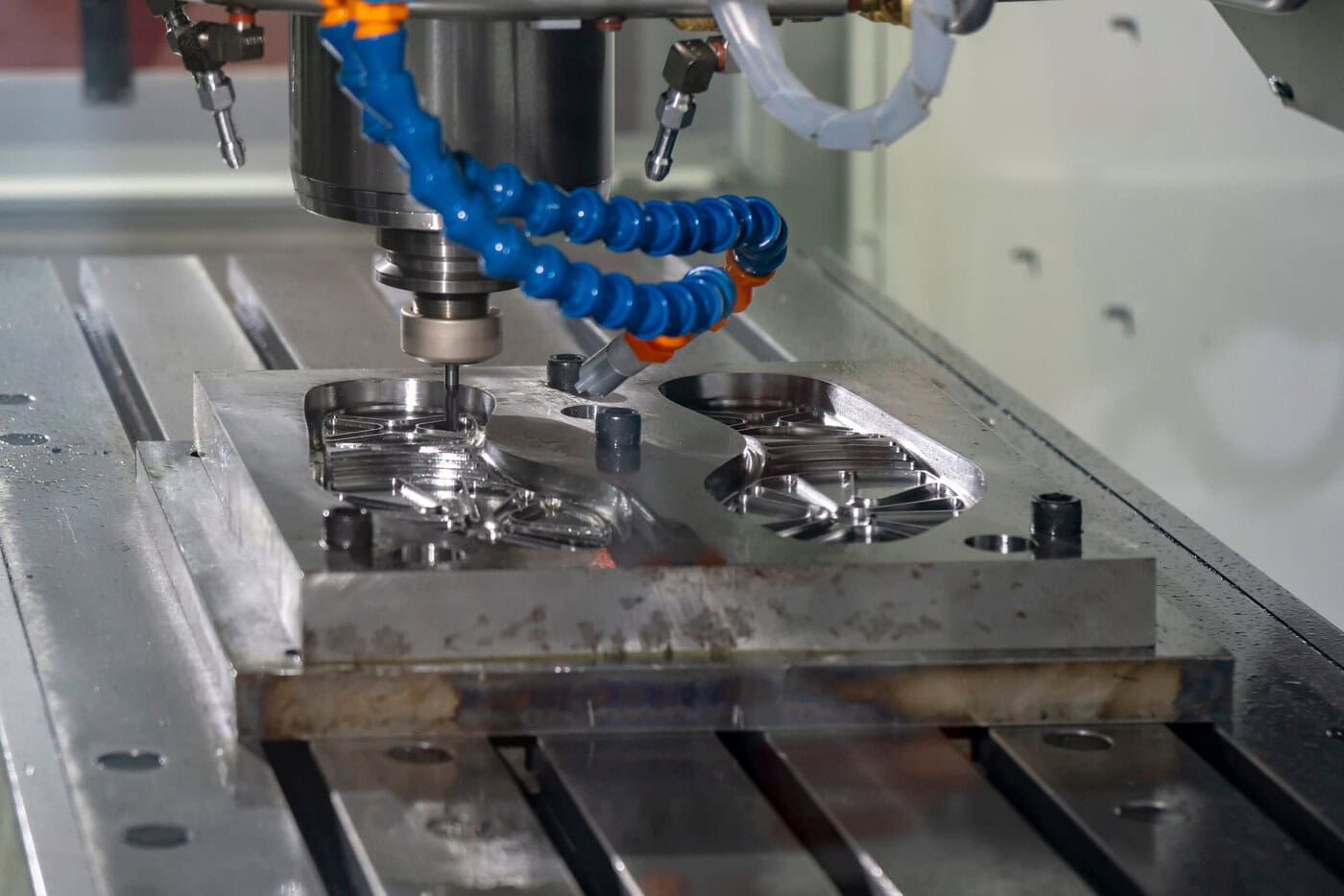 Close-up of a CNC machine in operation, carving intricate designs into a metal piece. The machine features a blue coolant hose, and shiny metal chips are visible, highlighting precision engineering and advanced manufacturing technology.