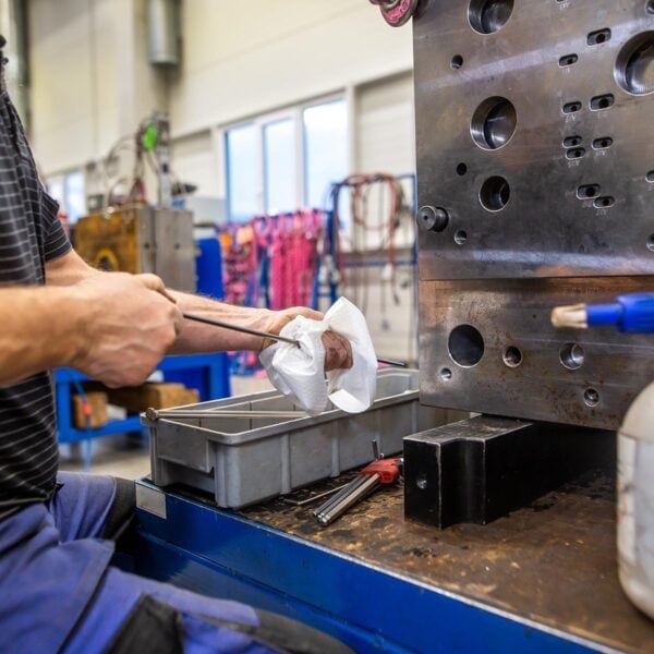 A Person Cleaning A Metal Tool In An Industrial Workshop. There Is A Large Metallic Machine With Holes On The Right And Various Tools And Equipment In The Background. The Person Is Using A Cloth And Holding A Long Metal Rod.