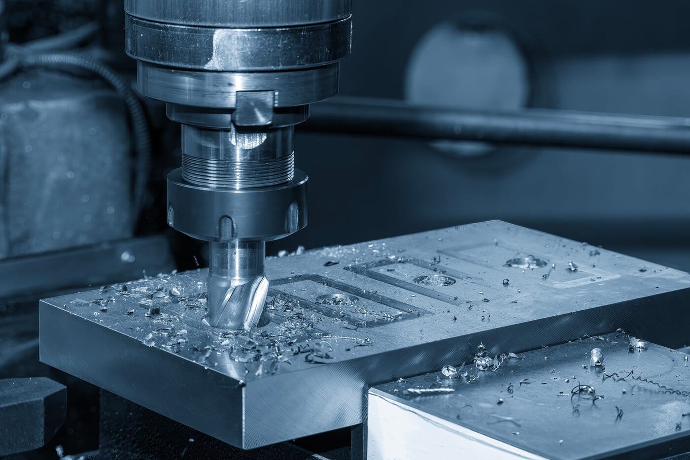 A CNC milling machine processes a metal workpiece, creating precise rectangular grooves. Metal shavings and debris are scattered around the area, highlighting the machining process. The image is tinted in a bluish hue.