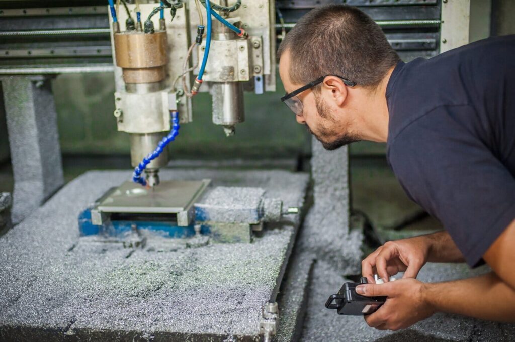 Amid A Whirlwind Of Metallic Shavings, A Person Wearing Safety Glasses Intensely Examines A Cnc Milling Machine In Operation. They Clutch A Tool, Fully Absorbed In The Milling Process, While Weighing The Cnc Machine Cost Against Its Precision And Efficiency.