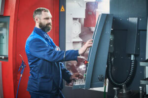 A Man With A Beard, Wearing A Blue Industrial Uniform, Operates A Large Cnc Machine In A Workshop. Focused On Pressing Buttons On The Control Panel, He Navigates The Complexities Of Machinery And Industrial Equipment, Mindful Of The Cnc Machine's Cost.