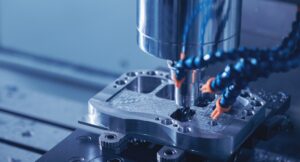 Close-Up Of A Cnc Milling Machine Working On A Metal Piece. The Sharp Cutting Tool Is Surrounded By Blue Tubes Spraying Coolant, Creating A Precise And Detailed Cut. The Metal Surface Has Visible Drill Marks And Shavings.