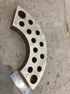 A Curved Metal Block With Multiple Circular Holes Arranged In A Grid Pattern. A Ruler Beside The Block Measures Its Length On A Concrete Surface.