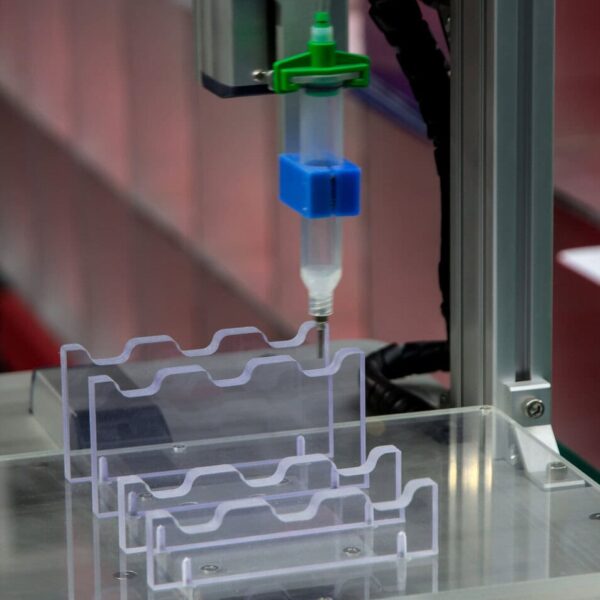 A Close-Up Of An Automated Machine, Possibly Part Of An Acrylic Cnc Machining Service, Dispensing Liquid Into A Clear Plastic Mold For Manufacturing Or Laboratory Use, With A Visible Nozzle And Colored Components.