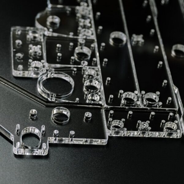 Close-Up Of Several Clear Panels With Various Holes And Cutouts, Arranged On A Dark Surface—Possibly Components Created Through An Acrylic Cnc Machining Service For Electronics Or A Custom Mechanical Assembly.