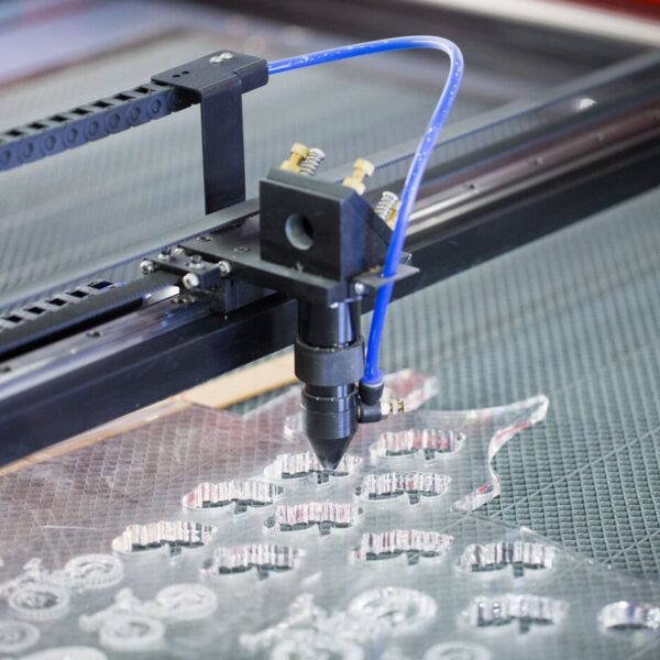 A Laser Cutting Machine With A Blue Tube Is Engraving And Cutting Intricate Shapes Out Of A Clear Acrylic Sheet On A Gridded Work Surface, Showcasing The Precision Of An Acrylic Cnc Machining Service.