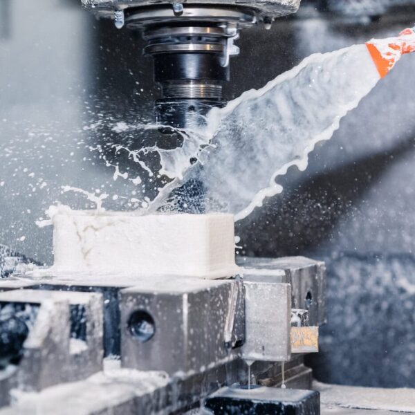 A Cnc Milling Machine From Our Abs Cnc Machining Service Carves A White Block While Coolant Fluid Is Sprayed Onto The Cutting Tool, Creating Splashes And Mist Inside The Enclosed Workspace.