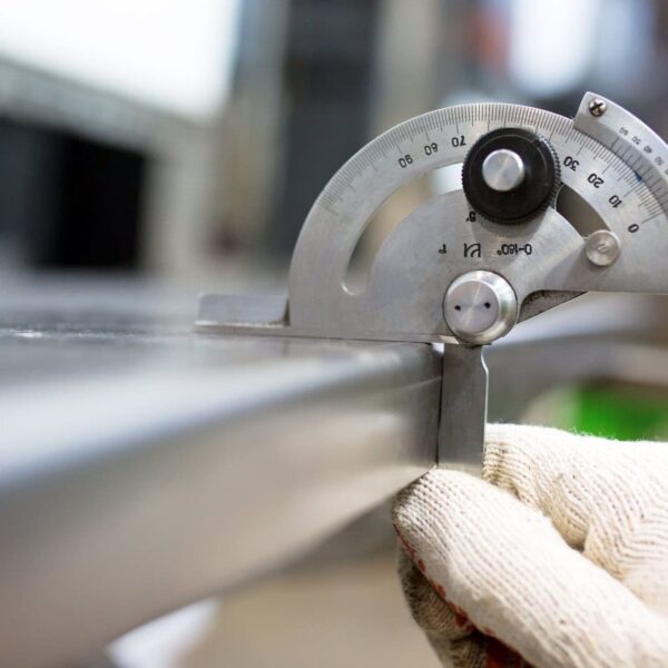 A Gloved Hand Holds A Metal Protractor Gauge, Measuring The Angle Of A Shiny Stainless Steel Sheet In An Industrial Or Workshop Setting. The Background Is Blurred.