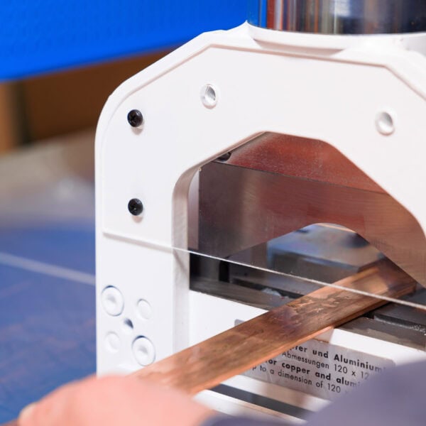 A Close-Up Of A Metalworking Machine Cutting A Rectangular Copper Bar, With A Person’s Hand Holding The Bar In Place. The Machine Has A White Exterior And The Workspace Has A Blue Surface.