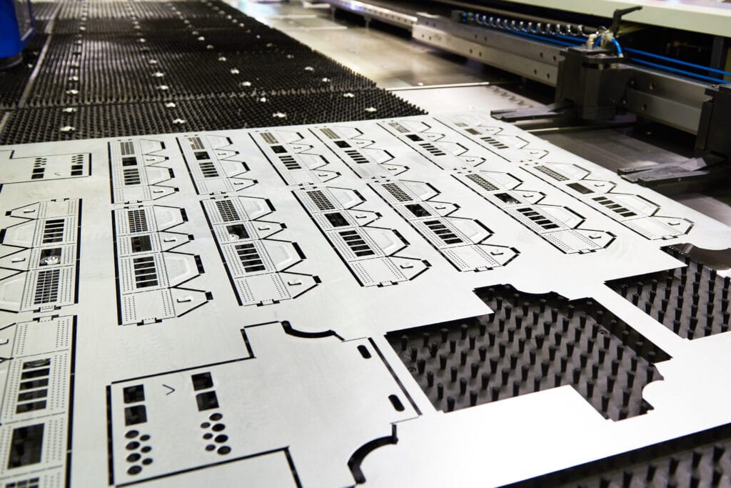 A close-up of a CNC machine cutting precise patterns into a large metal sheet, with intricate geometric shapes and detailed cutouts visible on the surface. The machine operates on a perforated black work surface.