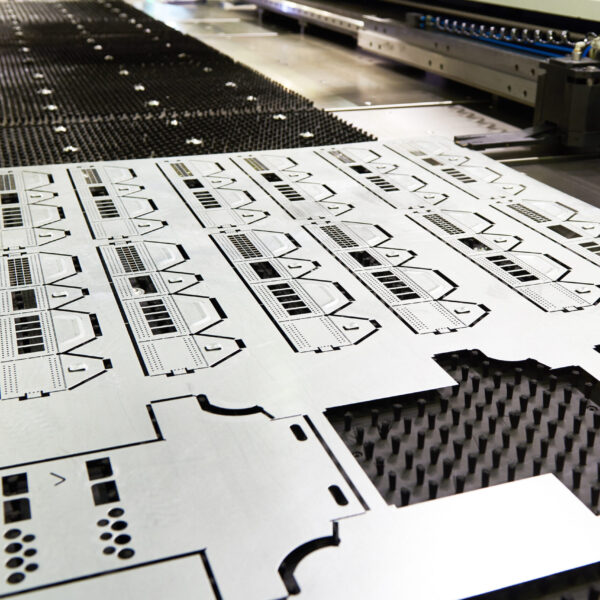 A Close-Up Of A Cnc Machine Cutting Precise Patterns Into A Large Metal Sheet, With Intricate Geometric Shapes And Detailed Cutouts Visible On The Surface. The Machine Operates On A Perforated Black Work Surface.