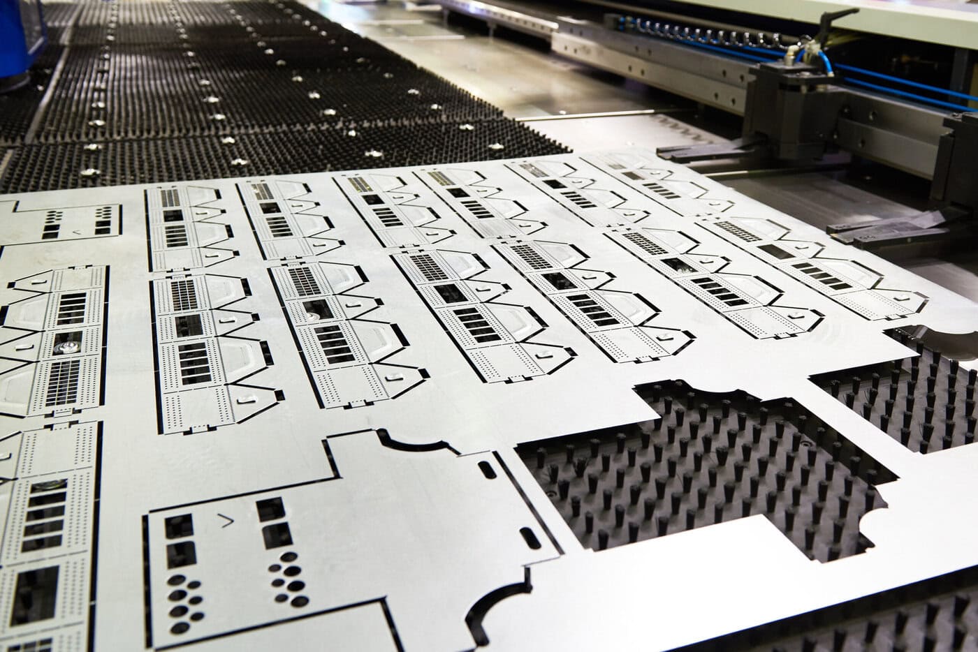 A sheet of metal with precise patterns cut out by a CNC machine, showing various geometric shapes and cutouts, placed on a perforated worktable in an industrial setting.