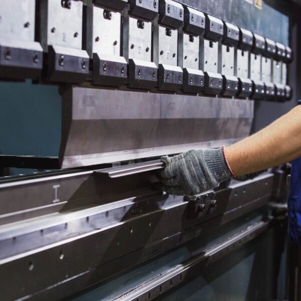 A Worker Wearing Gloves And A Blue Uniform Operates An Industrial Metal Bending Machine, Positioning A Metal Sheet Under The Machine’s Press In A Factory Setting.