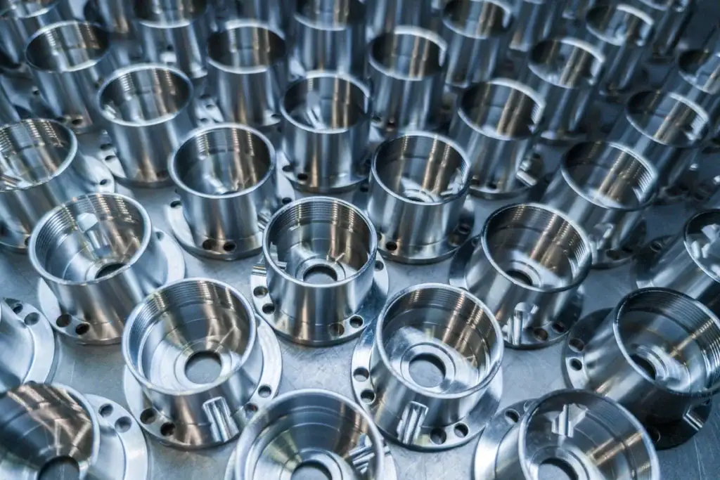 Rows Of Shiny, Metallic Cylindrical Machine Parts With Threaded Interiors Are Neatly Arranged In A Grid Pattern On A Reflective Surface, Showing Precise Manufacturing And Uniformity.