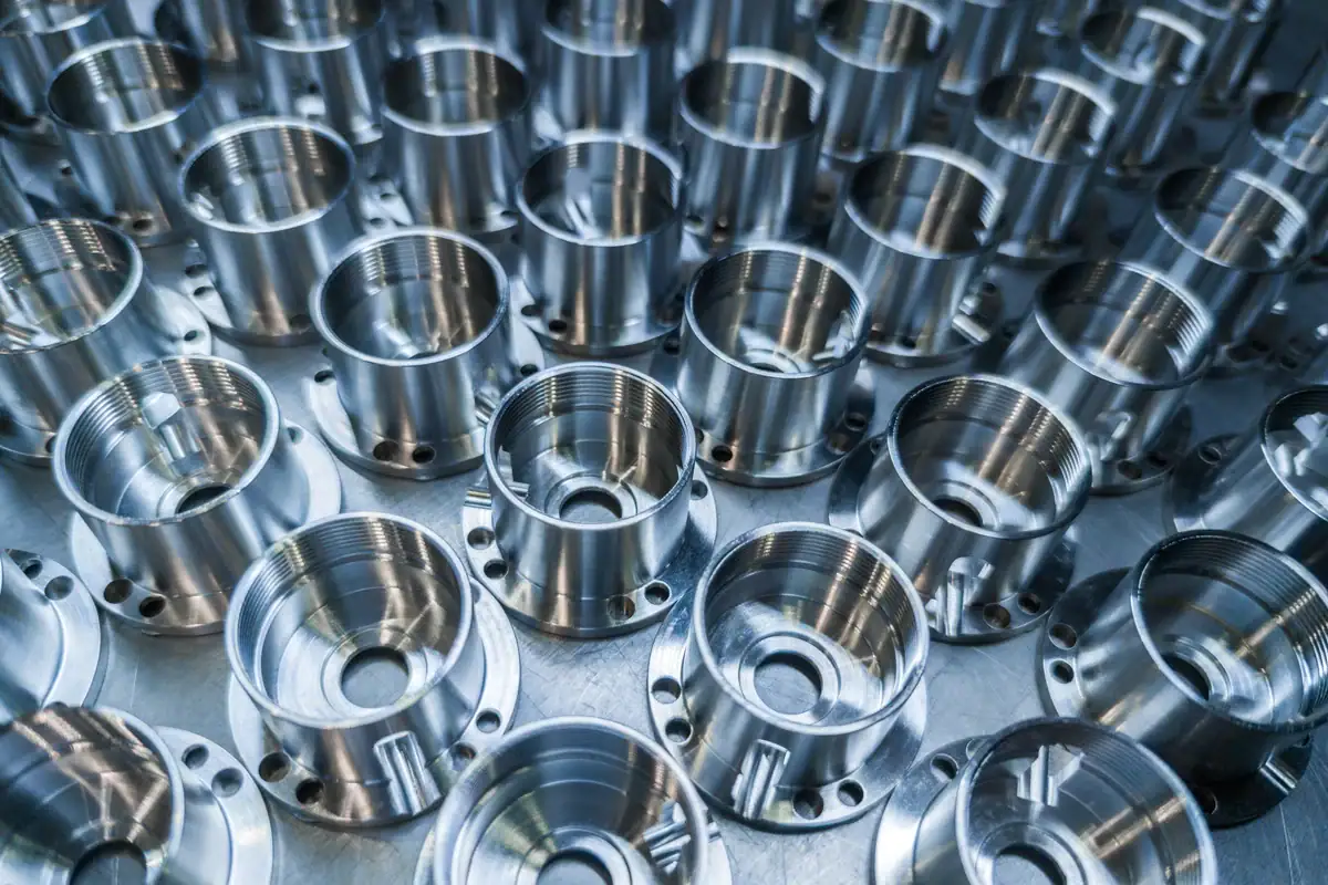 Rows of shiny, metallic cylindrical machine parts with threaded interiors are neatly arranged in a grid pattern on a reflective surface, showing precise manufacturing and uniformity.
