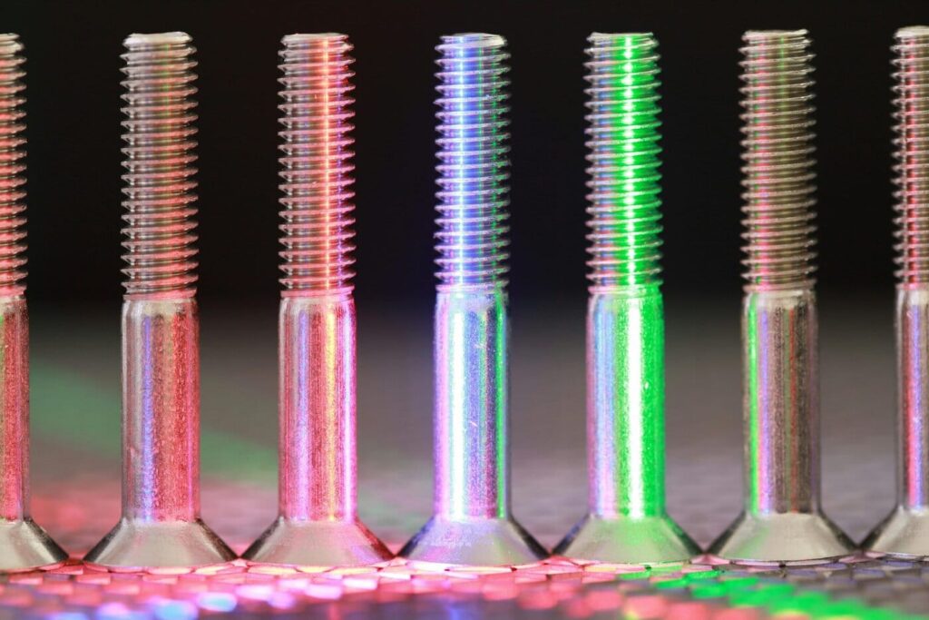 A Close-Up Of Several Metal Screws, Finished In Anodized Titanium Colors, Standing Upright In A Row And Illuminated By Red And Green Lights That Create Colorful Reflections On A Textured Surface.