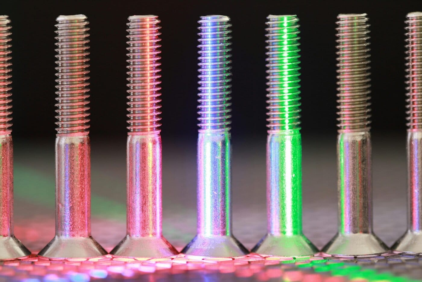 A close-up of several metal screws, finished in anodized titanium colors, standing upright in a row and illuminated by red and green lights that create colorful reflections on a textured surface.