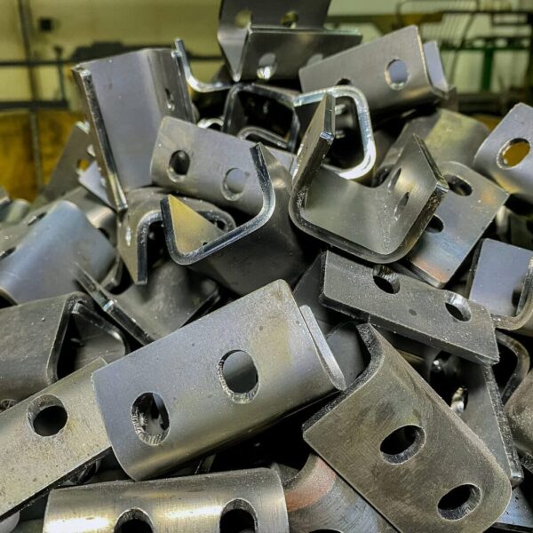 A Close-Up Of A Pile Of Shiny, Custom Metal Brackets With Holes, Stacked Together In An Industrial Setting. The Brackets Have A Bent Shape And Appear Freshly Manufactured.