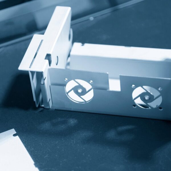 A Close-Up Of A Partially Assembled Metal Custom Computer Enclosure With Circular Fan Cutouts On One Side, Placed On A Dark Work Surface. The Image Has A Blue Tint, Emphasizing An Industrial Or Technical Setting.