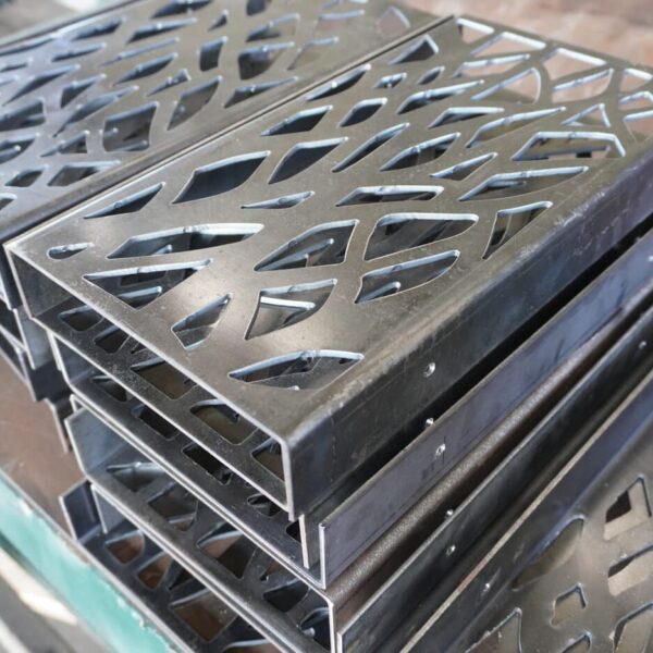 Stack Of Black Metal Sheets With Geometric, Leaf-Like Cutout Patterns, Likely Intended For Custom Sheet Metal Enclosures, Placed On A Workbench In An Industrial Or Workshop Setting.