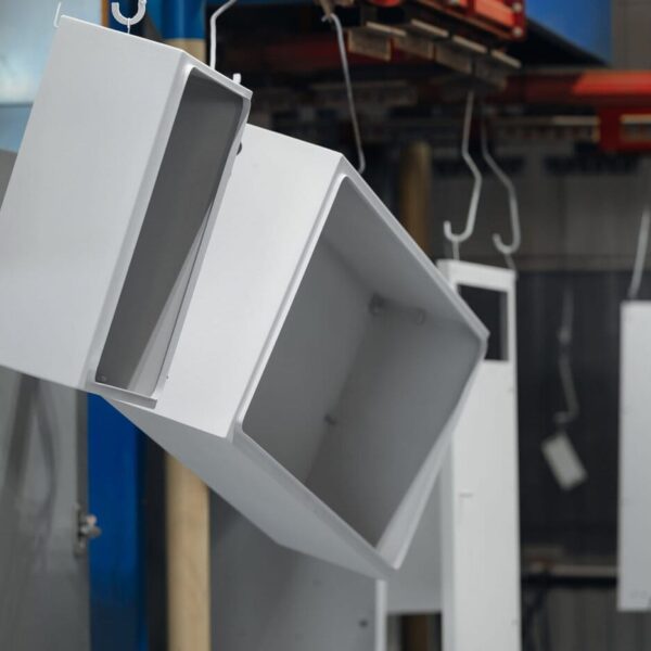 A Custom Sheet Metal Enclosure Is Suspended From A Hook In An Industrial Setting, Likely In A Paint Or Coating Booth. Other Similar Objects Hang In The Background On Metal Rails.