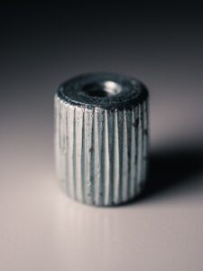 A Close-Up Photo Of A Small, Metallic, Cylindrical Object With Vertical Ridges And Straight Knurling, Featuring A Hole Through The Center And Resting On A Smooth, Light Surface.